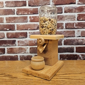 May include: A wooden nut dispenser with a glass jar filled with peanuts. The dispenser has a crank and a small wooden bowl to catch the nuts.