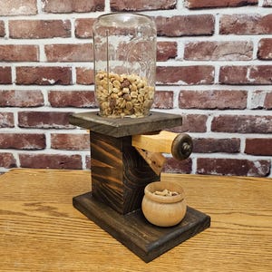 May include: A wooden nut dispenser with a glass jar filled with peanuts. The dispenser has a crank to dispense the nuts into a small wooden bowl.