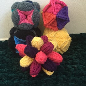 May include: Four crocheted balls in various colours and shapes. The balls are made of yarn in shades of pink, purple, yellow, orange, grey, and blue. The balls are arranged on a green rug.