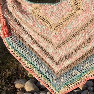 May include: A crocheted shawl with a colourful striped pattern in shades of pink, beige, blue, and green. The shawl has a fringe on the bottom edge.