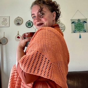 Puede incluir: Una mujer lleva una chal de ganchillo de color naranja quemado con un patrón de encaje. Está sosteniendo una taza en su mano derecha y mirando a la izquierda de la cámara.