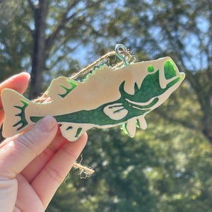 May include: A fish-shaped ornament featuring a cream-colored base with a dark green, stylized pattern across the body and fins. The ornament includes a silver-toned metal eyelet at the top and a piece of brown twine for hanging purposes.