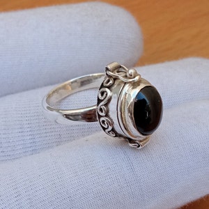 May include: A silver ring with a black stone and a hinged lid. The ring is decorated with a swirling design.