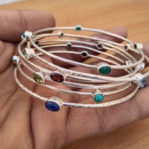 Puede incluir: Un conjunto de seis pulseras de plata con piedras preciosas de colores. Las pulseras están apiladas una encima de la otra y presentan una variedad de piedras preciosas, incluyendo turquesa, lapislázuli, granate, peridoto y ónix.