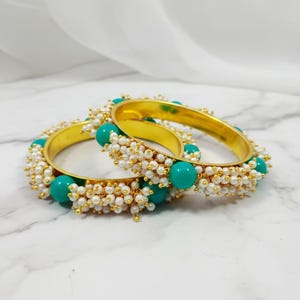 Op de afbeelding: Twee gouden armbanden met turquoise kralen en kleine witte parels. De armbanden zijn versierd met een patroon van turquoise kralen en kleine witte parels. De armbanden liggen op een wit marmeren oppervlak.