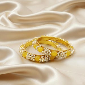 Op de afbeelding: Twee gouden armbanden met gele en witte kralenaccenten. De armbanden zijn versierd met clusters van kleine witte kralen en grotere gele kralen. De sieraden liggen op een satijnen stof.