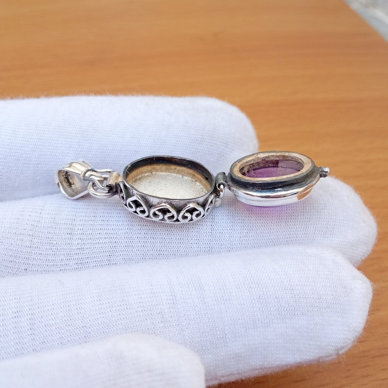 May include: A silver locket pendant with an ornate design and a hinged lid. The locket is open, revealing a purple gemstone inside.