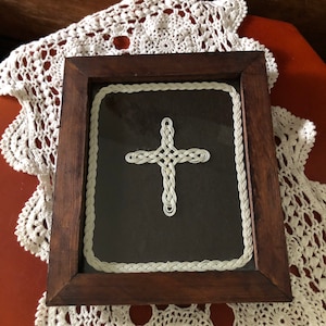 May include: A framed cross made of white braided rope on a black background. The frame is made of dark wood.