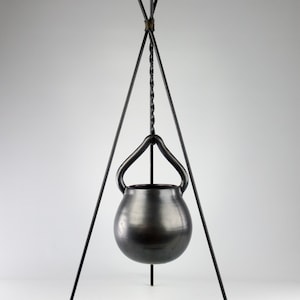 May include: A black ceramic pot hangs from a black metal tripod stand. The pot has a rounded shape and a wide opening. The stand is made of three legs that are connected at the top.