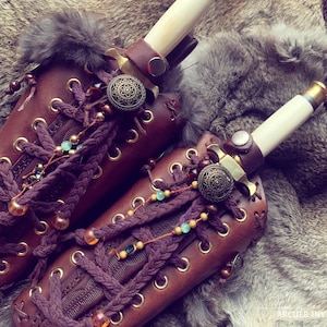 May include: Two brown leather arm bracers with decorative metal accents and beads. Each bracer has a dagger strapped to it with a white handle and gold accents.