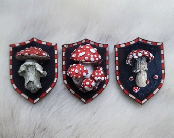 May include: Three red and white mushroom shaped ornaments with white spots are displayed in black and red shield-shaped frames. The frames have a white and red checkered pattern.