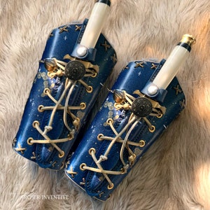 Puede incluir: Dos fundas de cuero azul con detalles blancos y dorados, diseñadas para guardar un cuchillo. Las fundas presentan un diseño de noche estrellada, costuras decorativas y un cierre de botón decorativo. El texto "Archer Inventive" es visible en la esquina inferior izquierda.