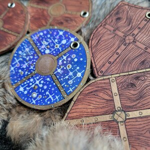 May include: Four leather shields with different designs. One shield is blue with white and gold accents and a circular pattern. The other three shields are brown with gold accents and a wood grain pattern.