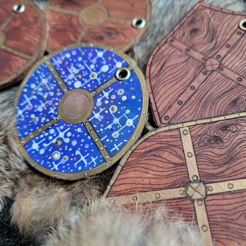 May include: Four leather shields with different designs. One shield is blue with white and gold accents and a circular pattern. The other three shields are brown with gold accents and a wood grain pattern.