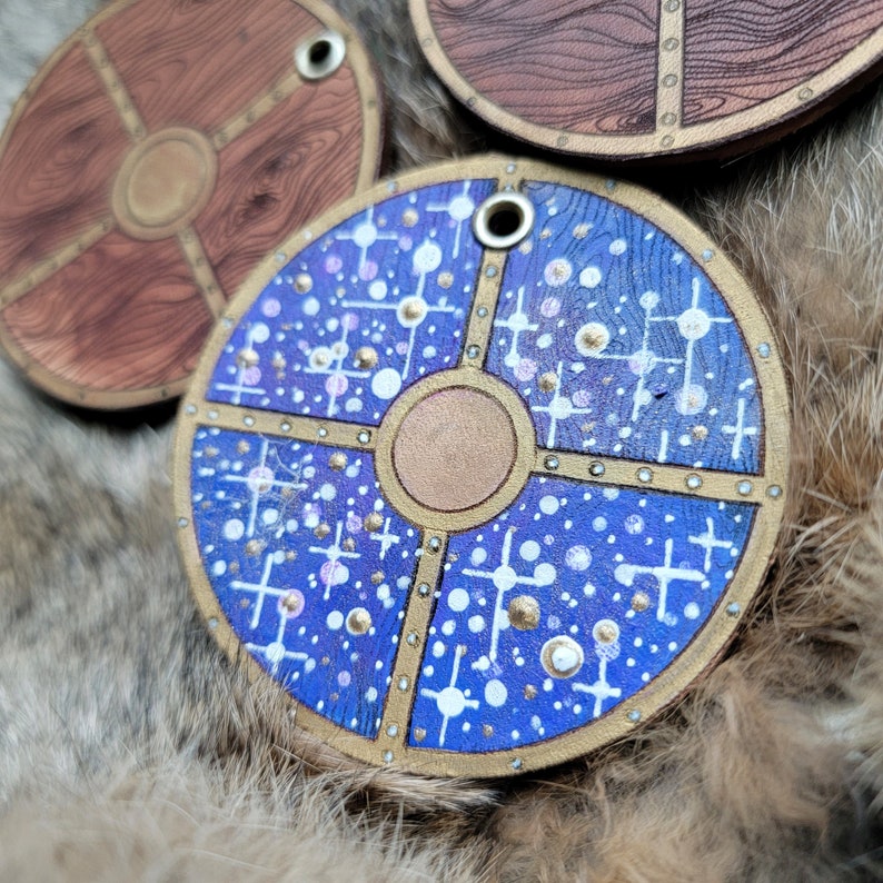 May include: A blue and gold wooden shield with a white and gold geometric pattern. The shield has a circular design with a central gold circle and a white and gold pattern around the edges.