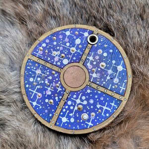 May include: A round, blue leather keychain with a gold design. The design features a circle in the center with a cross pattern around it. The keychain has a small hole at the top for attaching to a key ring.