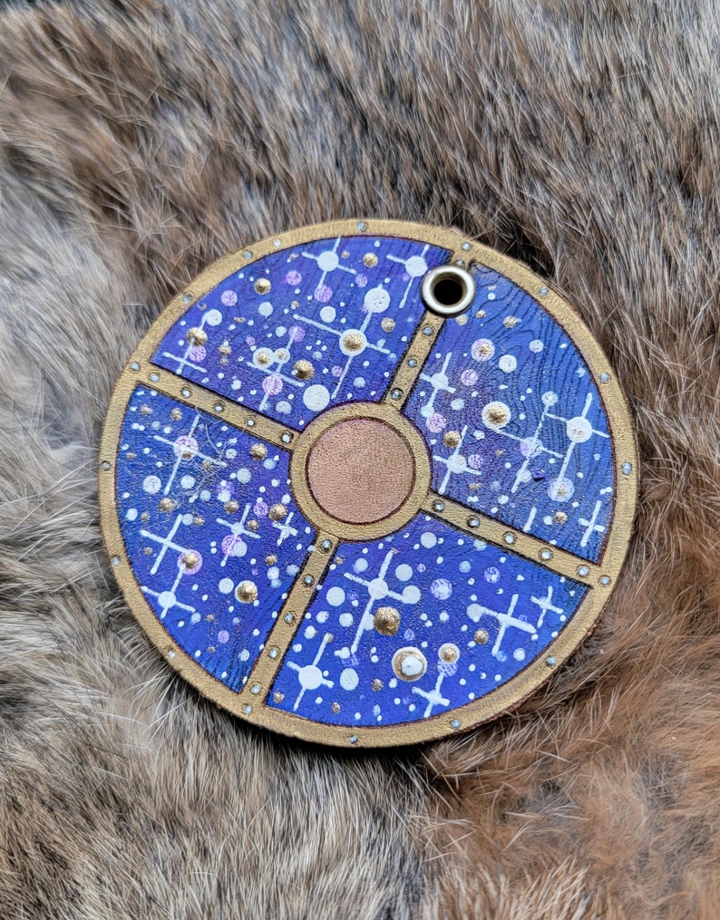 May include: A round, blue leather keychain with a gold design. The design features a circle in the center with a cross pattern around it. The keychain has a small hole at the top for attaching to a key ring.