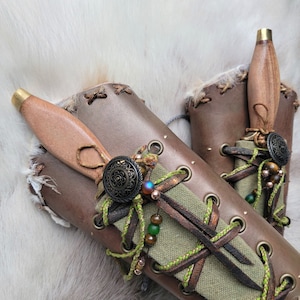 May include: Two leather sheaths with straps and buckles, each holding a wooden-handled dagger with a metal pommel. The sheaths are decorated with green and brown cords and beads.