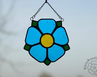 May include: A stained glass window hanging with a blue forget-me-not flower design with a yellow center. The flower is surrounded by green leaves.