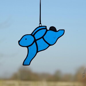May include: A blue stained glass bunny rabbit with black outlines, hanging from a string. The bunny is in a leaping pose with its ears back.