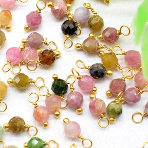May include: A collection of faceted tourmaline gemstone beads in various colors, including pink, green, brown, and yellow. Each bead is strung on a gold-colored wire with a loop at the top, suitable for jewelry making or crafting.