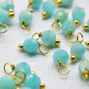 May include: A collection of turquoise blue gemstone beads with gold accents. Each bead has a small gold loop for attaching to jewelry.