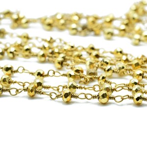 May include: Close-up of several strands of gold-colored beaded chain. The beads have a faceted, geometric shape and are strung together with delicate gold-toned wire and chain links. The background is white.
