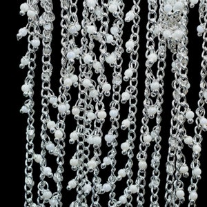 May include: A silver chain necklace with white beads hanging from it. The necklace is on a black background.