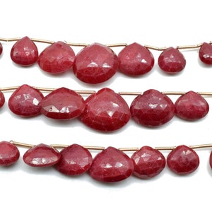 Pode incluir: Close-up de três fios de contas facetadas em forma de gota, de cor vermelho escuro. As contas são enfiadas em um fio fino, dourado. As superfícies facetadas das contas criam um efeito brilhante.