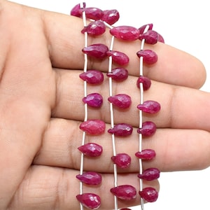 May include: A strand of ruby red gemstone beads in a teardrop shape. The beads are strung on a silver wire.