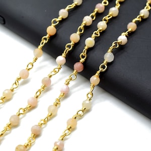 May include: Four strands of gold chain with pink and white beads. The chain is lying on a black surface.