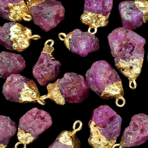 Op de afbeelding: Een verzameling van 20 ruwe robijn edelsteen hangers met vergulde accenten. De robijnstenen zijn diep rood van kleur en hebben een natuurlijke, onregelmatige vorm. De gouden accenten zijn een glanzend, metallic goud.