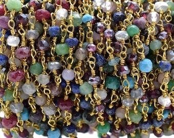May include: A close-up of a gold chain with various coloured gemstone beads. The beads are a mix of turquoise, green, red, purple, blue, and clear stones. The chain is made of small, linked gold rings.