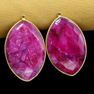 May include: Two oval-shaped ruby gemstone pendants with gold-toned metal settings. The gemstones are a deep pink color and have a faceted cut.