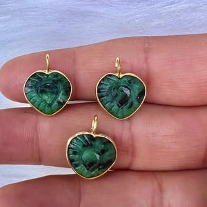 May include: Three heart-shaped pendants with a carved design and a vibrant green hue. Each pendant is encased in a gold-coloured setting with a small loop at the top for hanging. The pendants are displayed against a soft, white background.