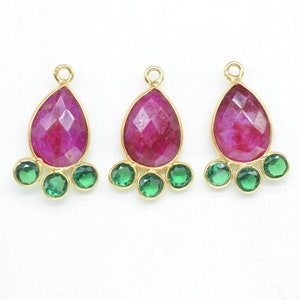 Op de afbeelding: Drie gouden druppelvormige hangers met roze edelstenen in het midden en drie kleinere groene edelstenen aan de onderkant van elke hanger.