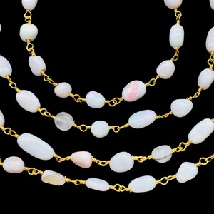 May include: Close-up of multiple strands of necklaces with white and pink oval-shaped beads. The beads are strung on delicate gold-coloured chains. The background is solid black.