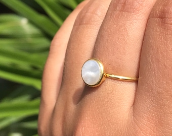 Könnte beinhalten: Ein goldener Ring mit einem weißen runden Edelstein. Der Ring befindet sich an einem Finger.