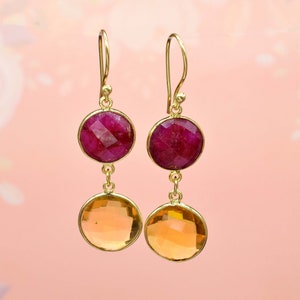 May include: Gold dangle earrings with two round gemstones. The top gemstone is a deep pink, and the bottom gemstone is a light orange.