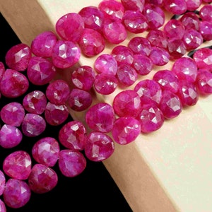 Pode incluir: Um fio de contas de rubi facetadas de cor rosa escura. As contas estão dispostas em fila sobre uma superfície rosa claro.