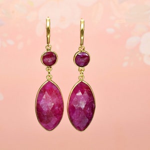 May include: A pair of gold earrings with two ruby gemstones. The larger gemstone is a teardrop shape and the smaller gemstone is round. The earrings are dangling.