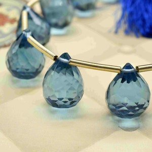 Könnte beinhalten: Nahaufnahme einer Kette aus facettierten, tropfenförmigen blauen Edelsteinen. Die Edelsteine sind auf einem goldfarbenen Draht aufgereiht. Im Hintergrund ist eine blaue Quaste zu sehen. Die Edelsteine werden auf einer cremefarbenen, gesteppten Oberfläche präsentiert.