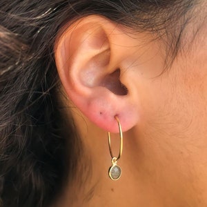 Peut inclure: Boucles d'oreilles créoles en or avec une petite pierre précieuse ronde grise suspendue au bas.