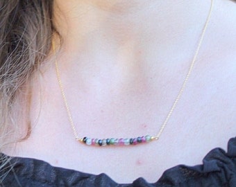 May include: A gold chain necklace with a bar of multi-colored gemstones. The gemstones are pink, green, and black.