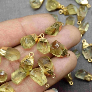 May include: A collection of raw, light green, crystal pendants with gold-toned jump rings. The pendants are various shapes and sizes, and are scattered on a hand.