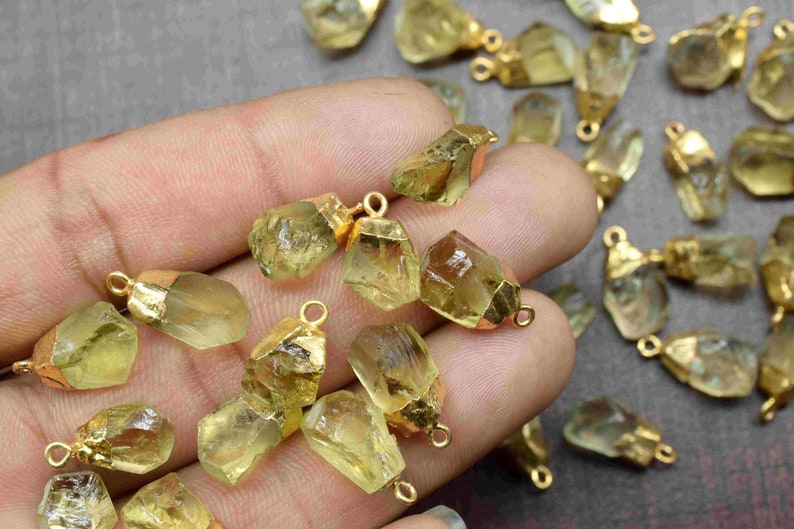 May include: A collection of raw, light green, crystal pendants with gold-toned jump rings. The pendants are various shapes and sizes, and are scattered on a hand.