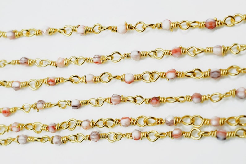 May include: Gold chain with pink and white beads. The chain is made of small gold links and the beads are round and have a marbled pattern.