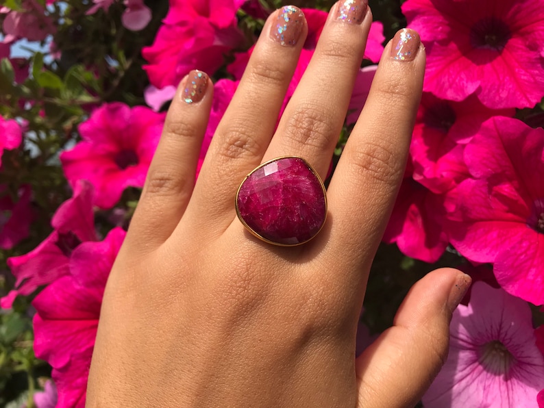 K&ouml;nnte beinhalten: Ein goldener Ring mit einem gro&szlig;en, facettierten, tief roten Edelstein. Der Ring wird an einer Hand mit rosafarbenem Nagellack getragen.