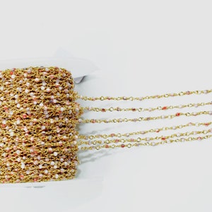 May include: A spool of gold chain with small pink and white beads. The chain is laid out in rows on a white surface.
