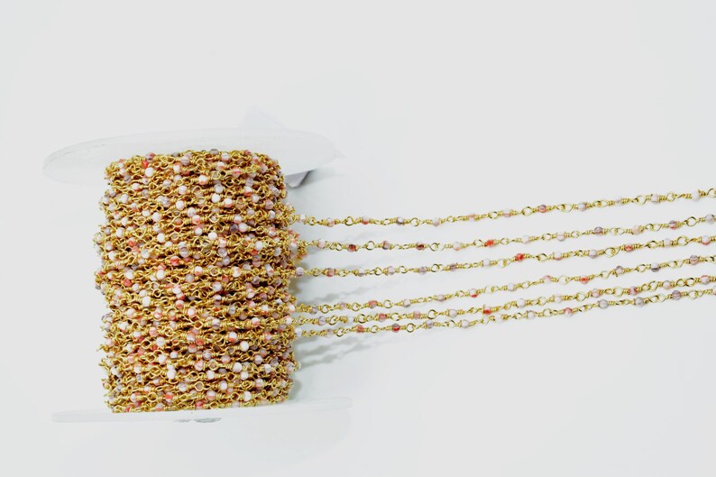May include: A spool of gold chain with small pink and white beads. The chain is laid out in rows on a white surface.